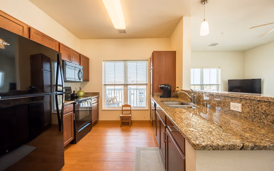 A modern kitchen with wooden cabinets, granite countertops, a black refrigerator, stove, microwave, and a coffee maker. There is a window with blinds at the end of the kitchen and a small wooden chair placed in front of it. The kitchen opens up to a living area with a TV visible in the background.