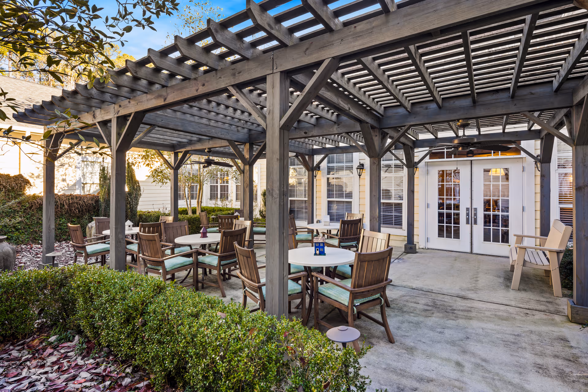 Outdoor patio with a wooden pergola covering several round tables and cushioned chairs in front of glass French doors.