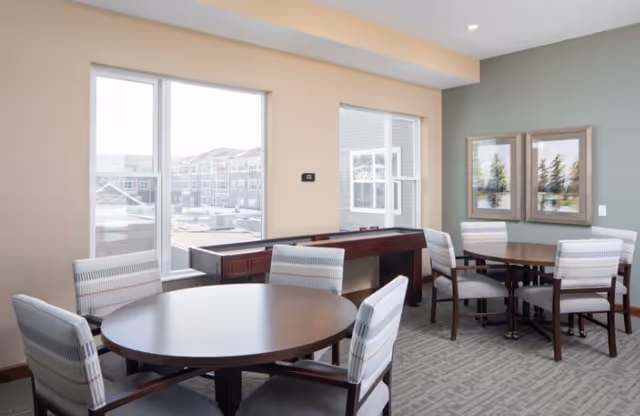 A bright room with two round wooden tables, each surrounded by four cushioned chairs with striped upholstery. The room has large windows letting in natural light, a shuffleboard table against the wall, and two framed landscape paintings on a green accent wall.