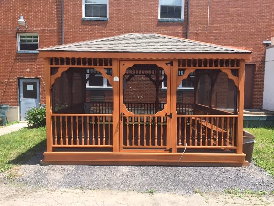 A wooden gazebo with a shingled roof and screened sides is situated outdoors on a gravel and grass area in front of a red brick building with several windows and a door.