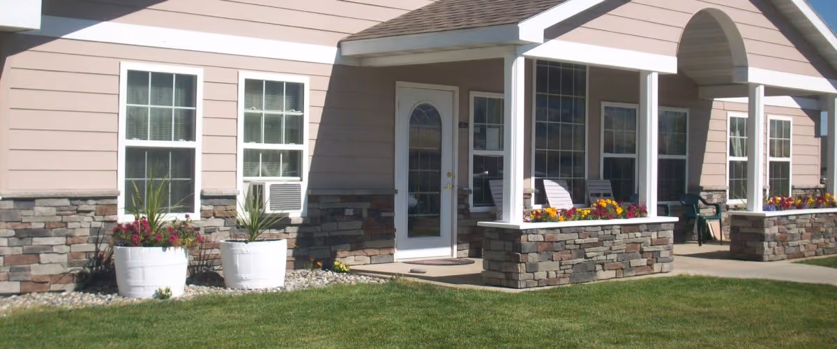 Exterior view of a building with beige siding and stone accents. The building has multiple windows, a white door with glass panels, and a covered porch area with white columns. There are flower pots with colorful flowers and green plants near the windows and on the porch. The lawn in front is green and well-maintained.