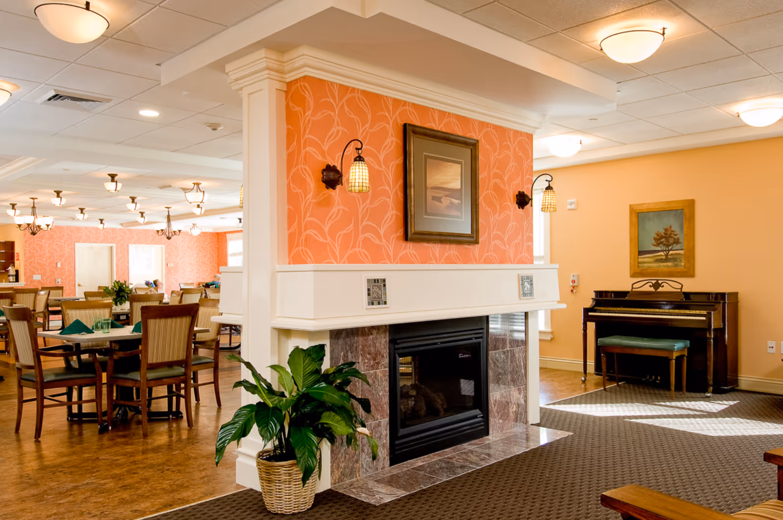A warm and inviting common area in a senior living facility featuring a central fireplace with a framed picture above it, a potted plant beside the fireplace, a piano with a bench against the wall, and several dining tables and chairs in the background. The walls are painted in soft orange and peach tones, and the room is well-lit with ceiling lights.