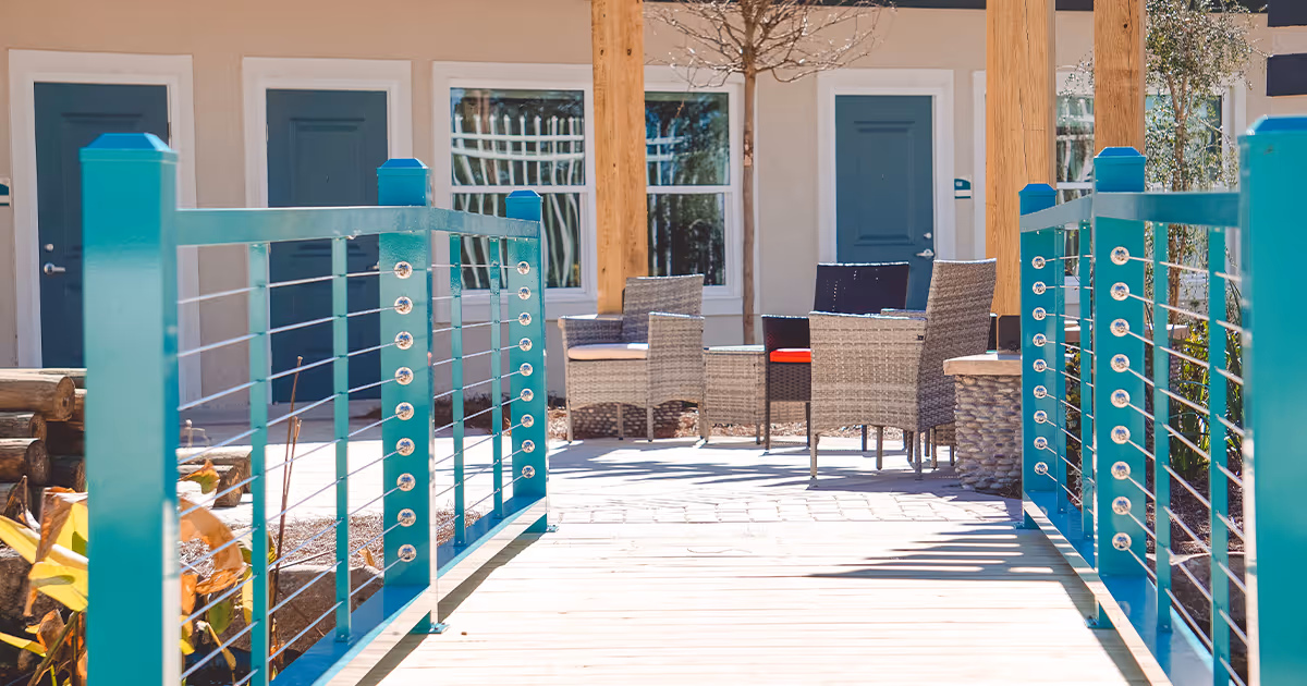 Outdoor patio area with a wooden walkway featuring teal railings leading to a seating area with wicker chairs and a table, set against a building with beige walls and blue doors and windows.