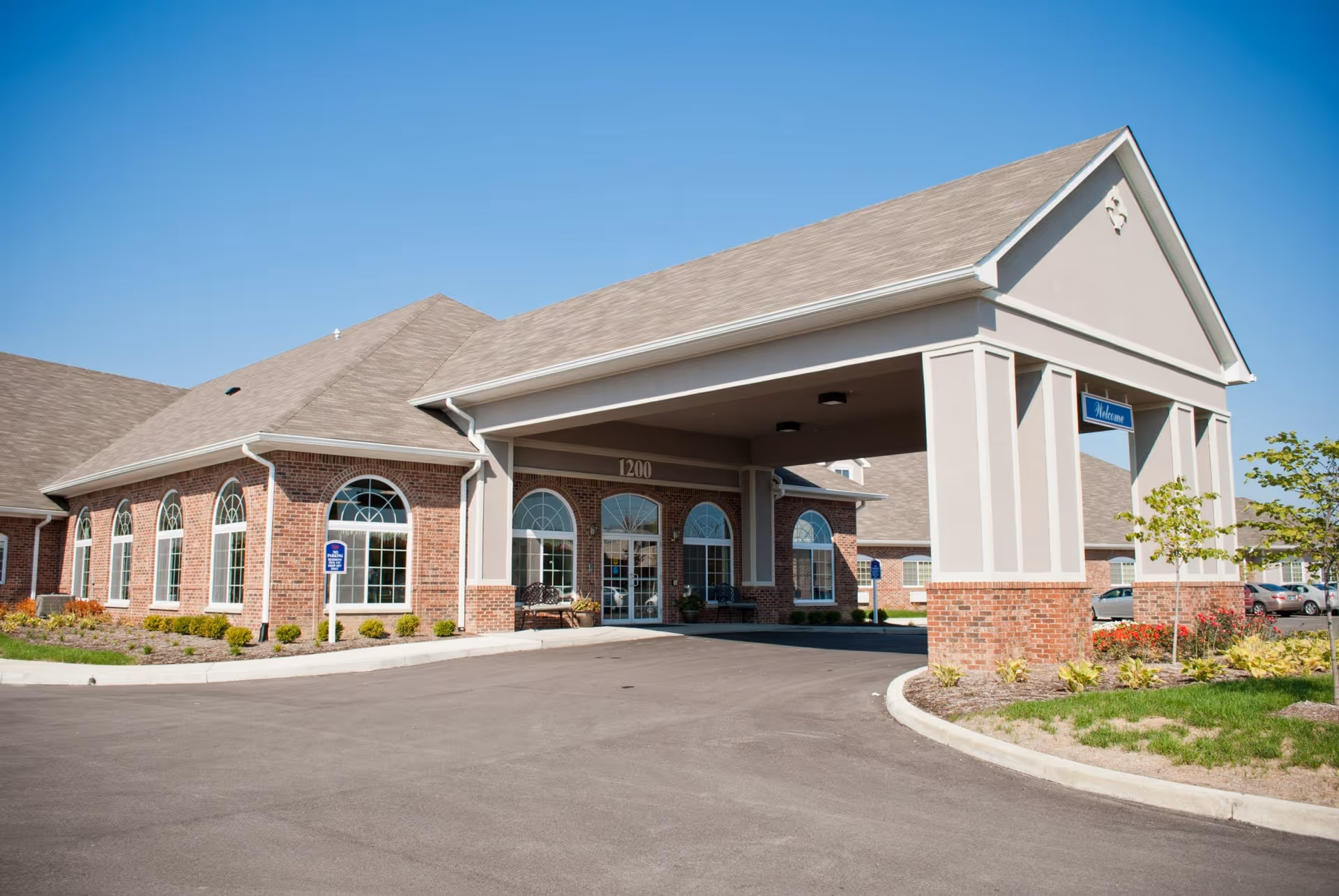 Exterior front entrance of a senior living facility with a covered drop-off area, brick and beige walls, arched windows, and a clear blue sky.