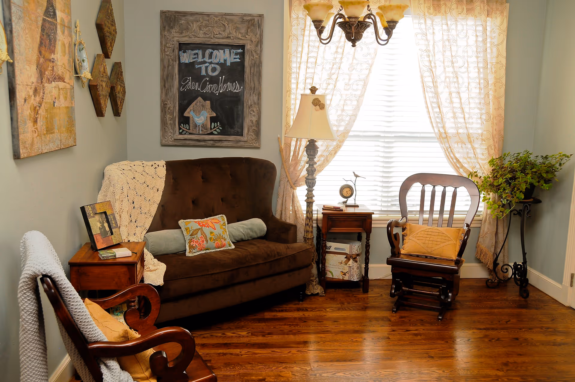 Cozy living room with a brown sofa, wooden chairs and side tables in front of a bright window with sheer curtains and a chalkboard sign reading "Welcome to Eden Care Homes".