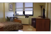 Small assisted-living bedroom with a single bed, window with blinds, a chair, dresser and a small table.