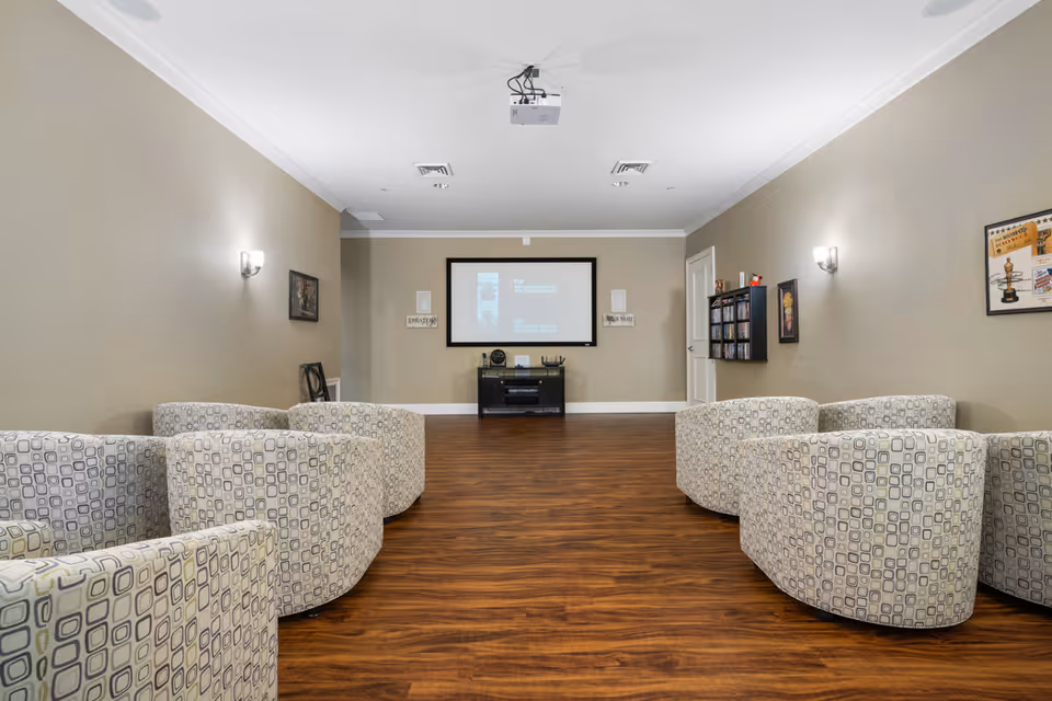 A media room with a projector screen on the far wall, a projector mounted on the ceiling, and six patterned armchairs arranged in two groups facing the screen. The room has wood flooring, beige walls with framed pictures, and wall-mounted lights.