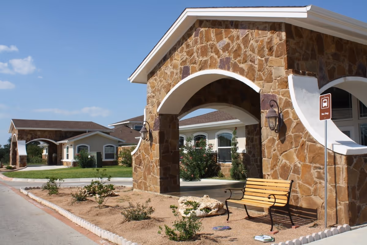 Outdoor view of a senior living facility with stone archways, a wooden bench, small plants, and a bus stop sign under a clear blue sky.