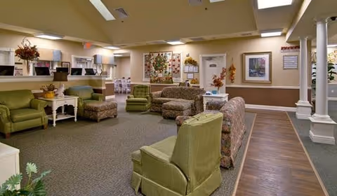 A spacious senior living facility common area with multiple upholstered chairs and sofas arranged for conversation. The room features a mix of green and patterned furniture, a carpeted floor with a wood floor section, decorative plants, framed artwork on the walls, and soft overhead lighting.