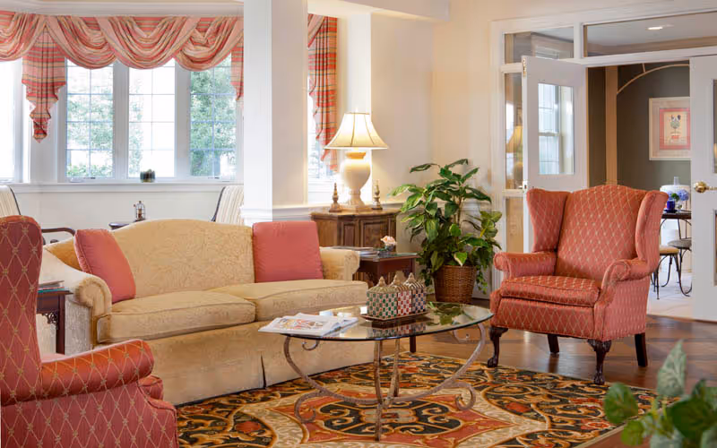 A cozy living room area with a beige sofa adorned with red cushions, two red patterned armchairs, a glass coffee table with decorative items and magazines, a colorful patterned rug, a wooden side table with a lamp, and a large green potted plant. The room has large windows with red and white striped valances and an open doorway leading to another room with a table and chairs.