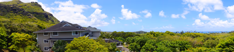 A multi-story building surrounded by lush green trees and vegetation under a bright blue sky with scattered white clouds. Hills are visible in the background.