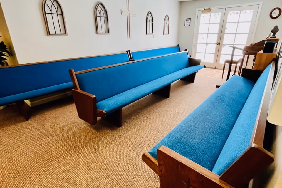 Small chapel-like room with rows of wooden pews upholstered in blue, wall crosses and glass doors at the far end.