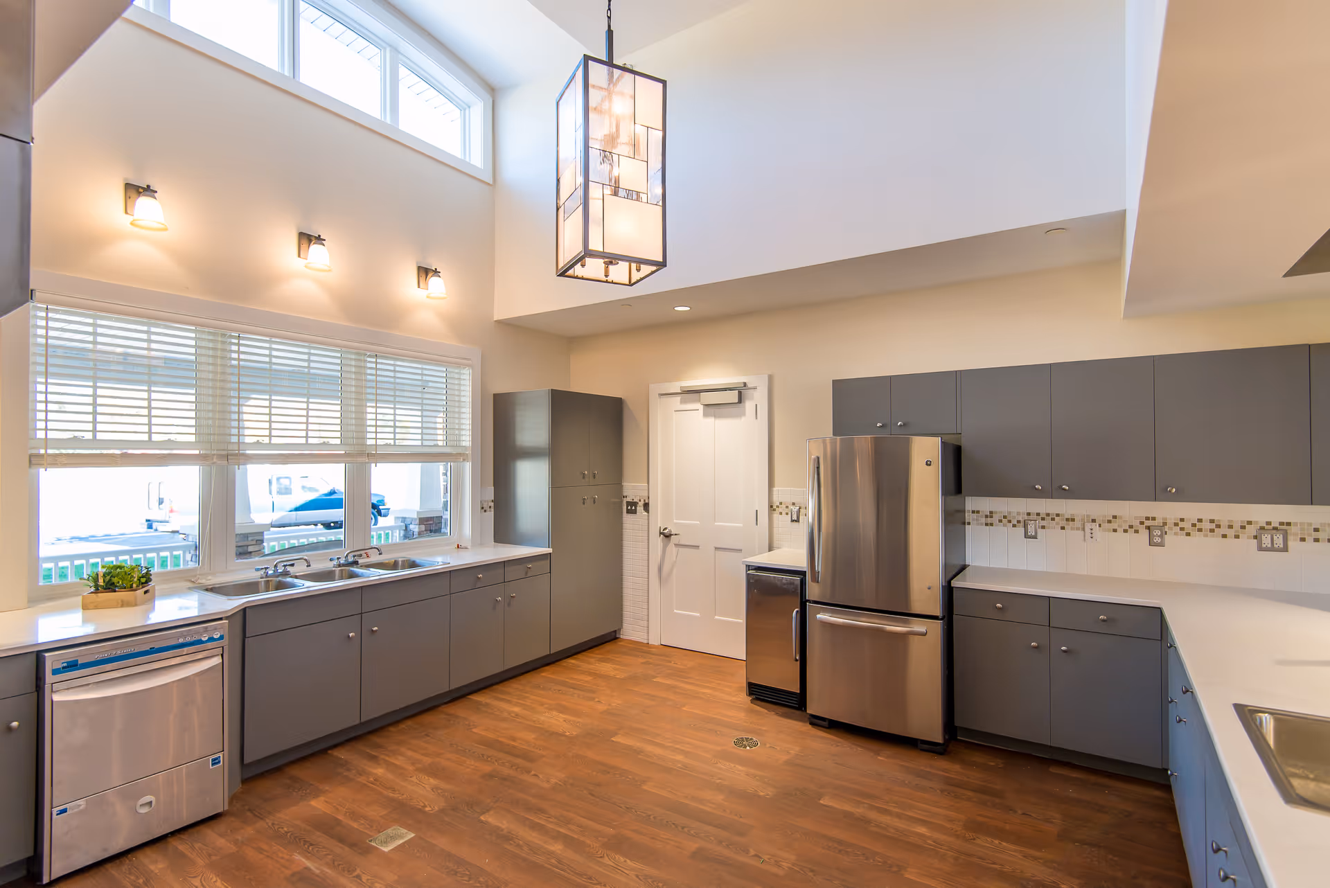 A modern kitchen with gray cabinets, stainless steel refrigerator and dishwasher, white countertops, wooden flooring, large windows with blinds, and a hanging rectangular light fixture.