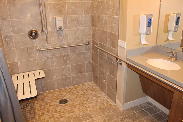 Accessible tiled bathroom with a roll-in shower, fold-down bench, grab bars, sink and wall-mounted soap dispensers.