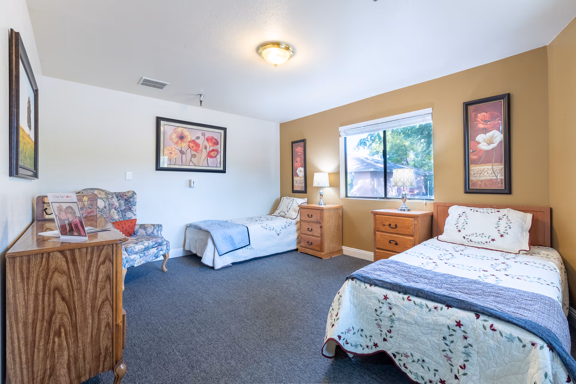 Bright memory-care bedroom with two twin beds, wooden nightstands, a floral armchair, and a window.