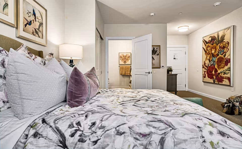 Bright, well-decorated bedroom with a patterned bed and pillows, bedside lamp, and an open door leading to a hallway with artwork.