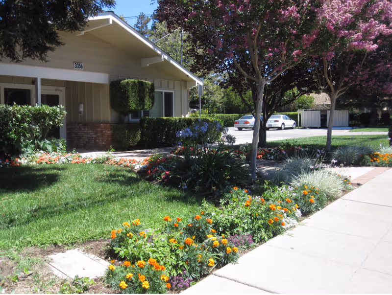 Exterior view of a single-story building with the number 3356, surrounded by a well-maintained garden with colorful flowers, green shrubs, and trees with pink blossoms. A sidewalk runs along the garden, and two parked cars are visible in the background.