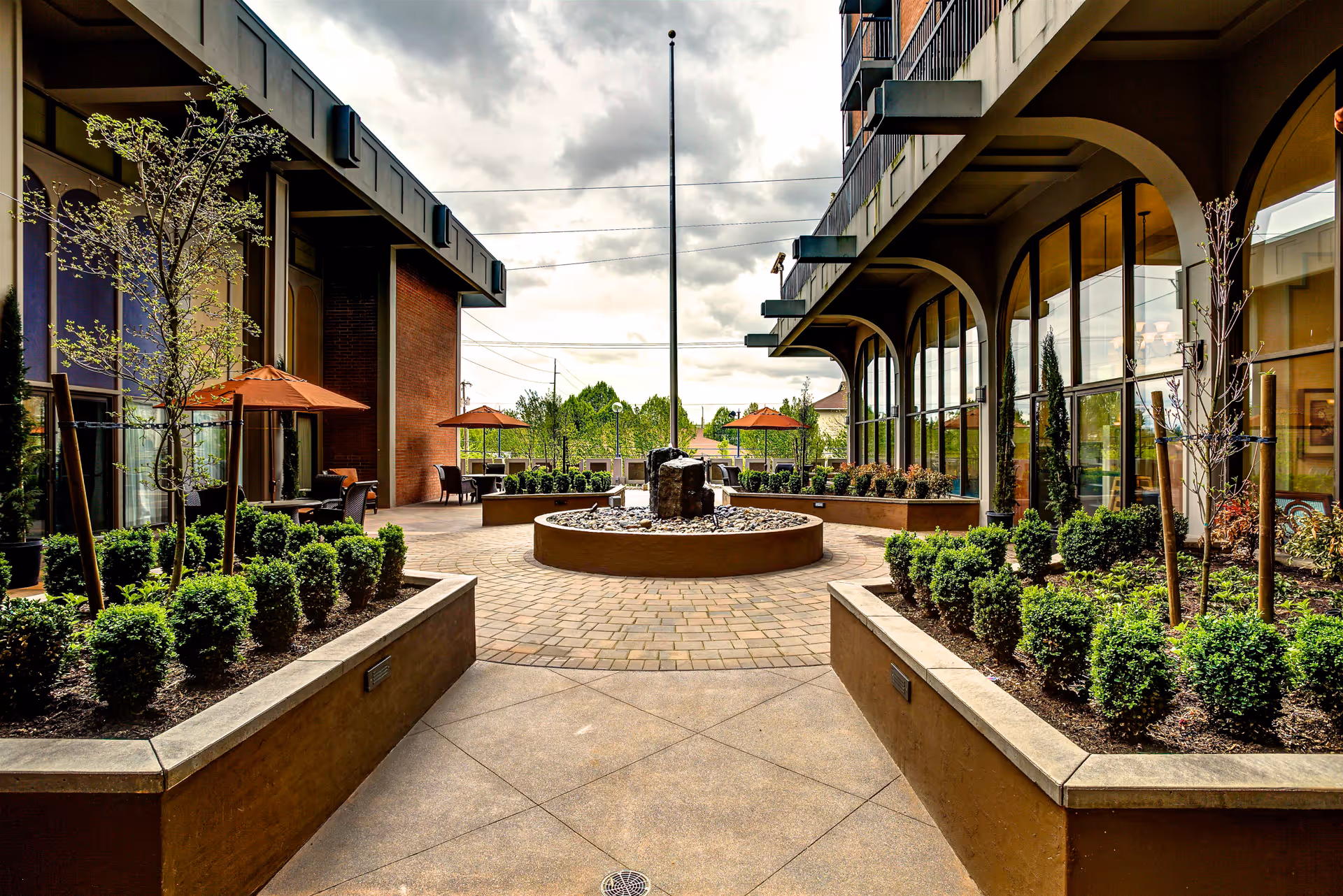 Outdoor courtyard area at Calaroga Terrace featuring a central circular stone water fountain surrounded by paved walkways, landscaped garden beds with small shrubs and young trees, and seating areas with tables and orange umbrellas. The courtyard is flanked by buildings with large windows and arched architectural details under a cloudy sky.