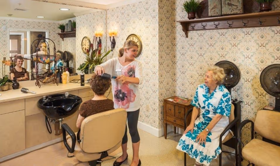 A hair salon area in a senior living facility where a woman is styling the hair of an elderly woman seated in a salon chair. Another elderly woman is sitting nearby, wearing a blue and white floral dress. The room has patterned wallpaper, a black salon sink, and various hair styling tools on the counter.