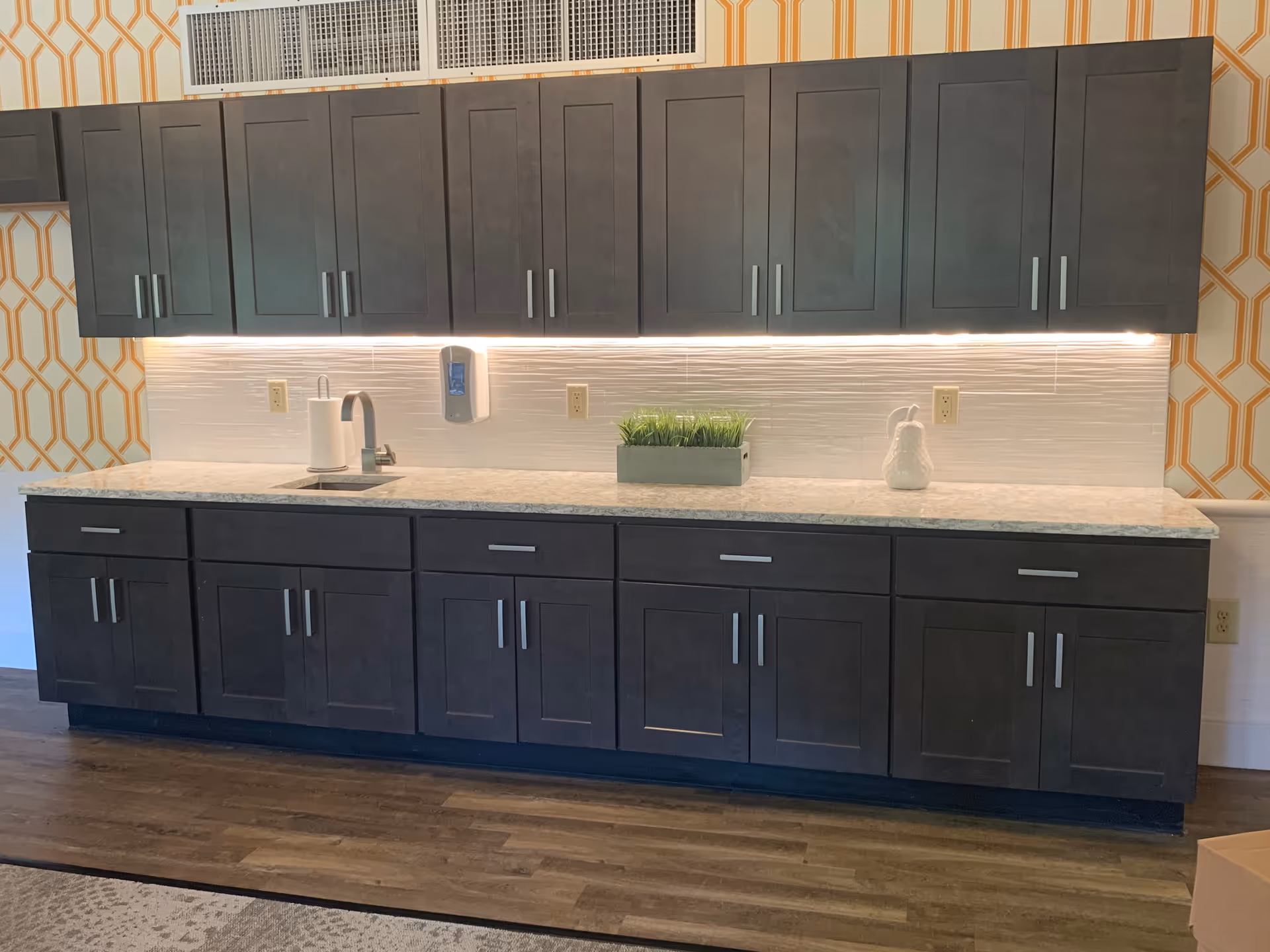 A modern kitchenette area with dark wood cabinets both above and below a light-colored marble countertop. The countertop features a small sink with a faucet, a paper towel holder, a rectangular planter with green grass, and a white decorative item. The backsplash is white with a textured pattern, and there is under-cabinet lighting illuminating the countertop. The wall behind the kitchenette has a yellow and white geometric patterned wallpaper, and the floor is a dark wood laminate.