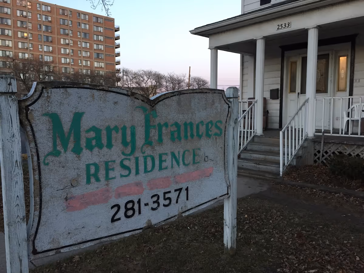 Worn wooden sign reading 'Mary Frances Residence' and a phone number in front of a small porch-style house with steps and columns.