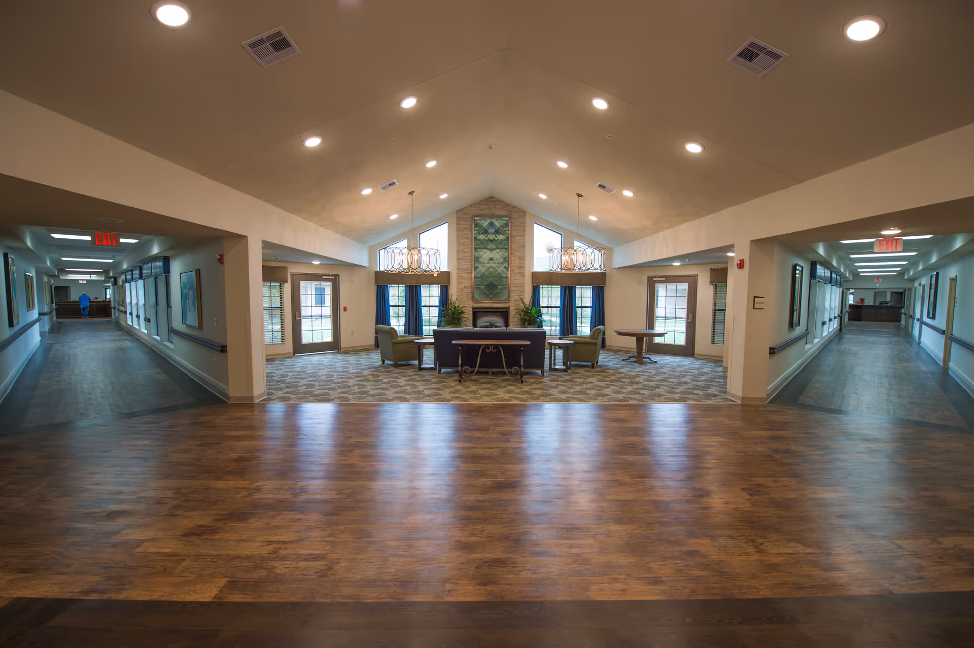 Spacious senior living common area with seating, chandeliers, and a fireplace, flanked by two long hallways.