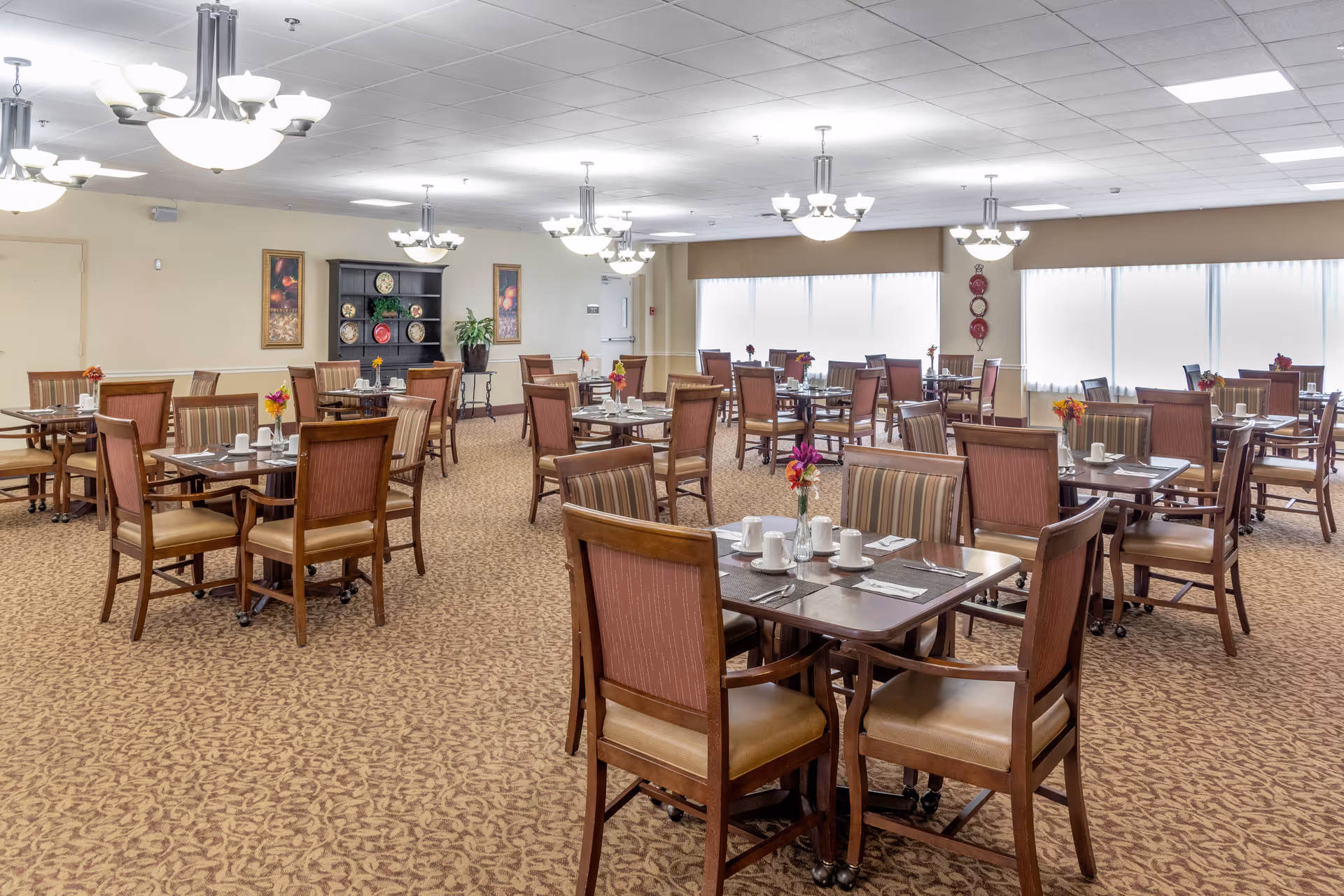 A spacious dining room with multiple wooden tables and chairs arranged neatly. Each table is set with white cups, saucers, and silverware, and decorated with small flower arrangements. The room has beige walls, carpeted floors with a patterned design, and several ceiling light fixtures providing bright illumination. Large windows with white blinds allow natural light to enter the room. There are framed pictures and a black cabinet with decorative plates on the far wall.