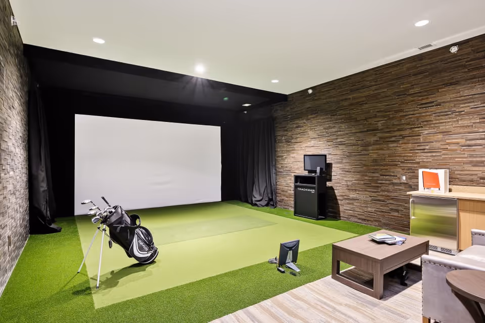 Indoor golf simulator room with artificial green turf, a golf bag with clubs, a large white projection screen, a small monitor on a stand, and a seating area with a coffee table and chairs. The walls have a textured stone finish.