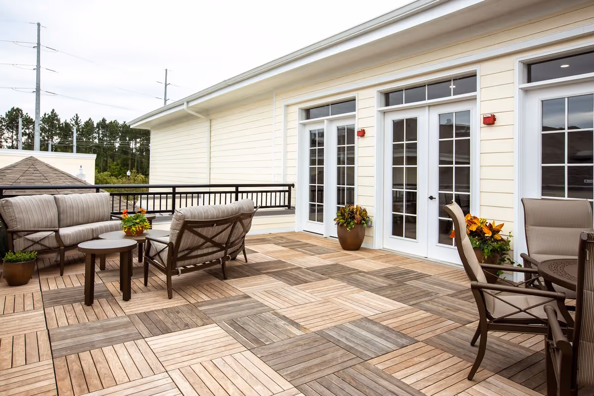 Outdoor patio area with wooden tile flooring, cushioned seating including a sofa and chairs, small round tables, potted plants, and large glass doors leading inside a light-colored building.