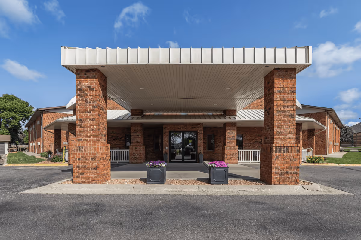 Front exterior view of a senior living facility with a covered entrance supported by brick pillars, two planters with purple flowers, and a clear blue sky in the background.