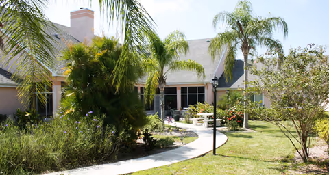 A sunny outdoor garden area at Brookdale Cape Coral featuring a paved walkway, palm trees, bushes, and a bench near the building with large windows and a chimney.