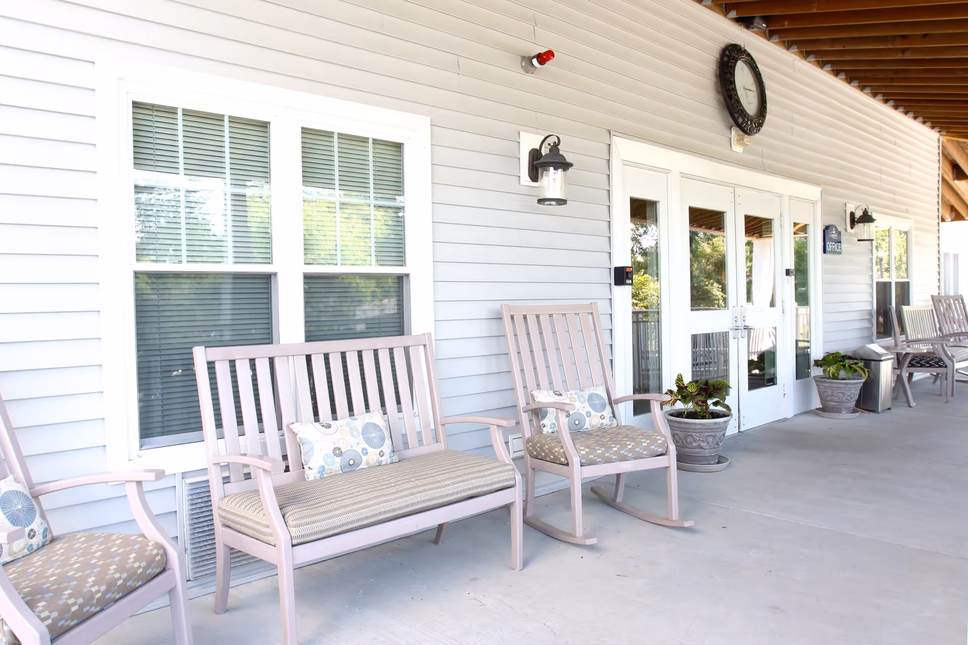 Outdoor covered porch area with light purple wooden rocking chairs and benches with patterned cushions and pillows. There are potted plants near the white double doors labeled 'Office' and windows with closed blinds on the light gray siding wall.