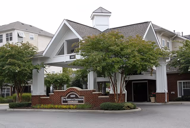 Entrance of The Reserve at Greenbrier facility featuring a covered drop-off area with white pillars and a peaked roof, surrounded by trees and landscaping.