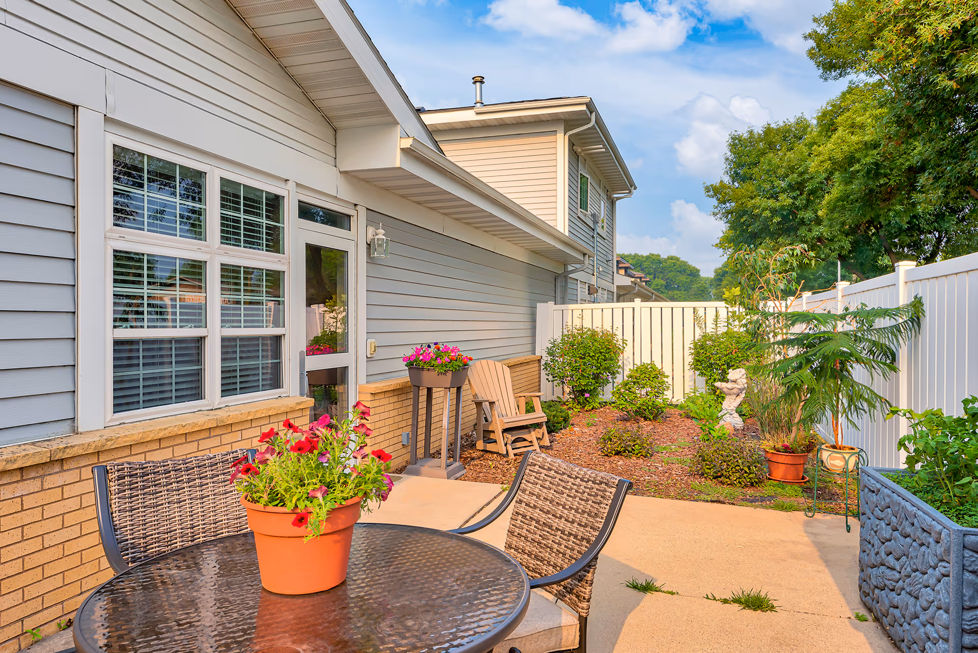 Sunny patio with a round glass table and potted flowers, wicker chairs, and a small landscaped garden beside a light-gray siding building.