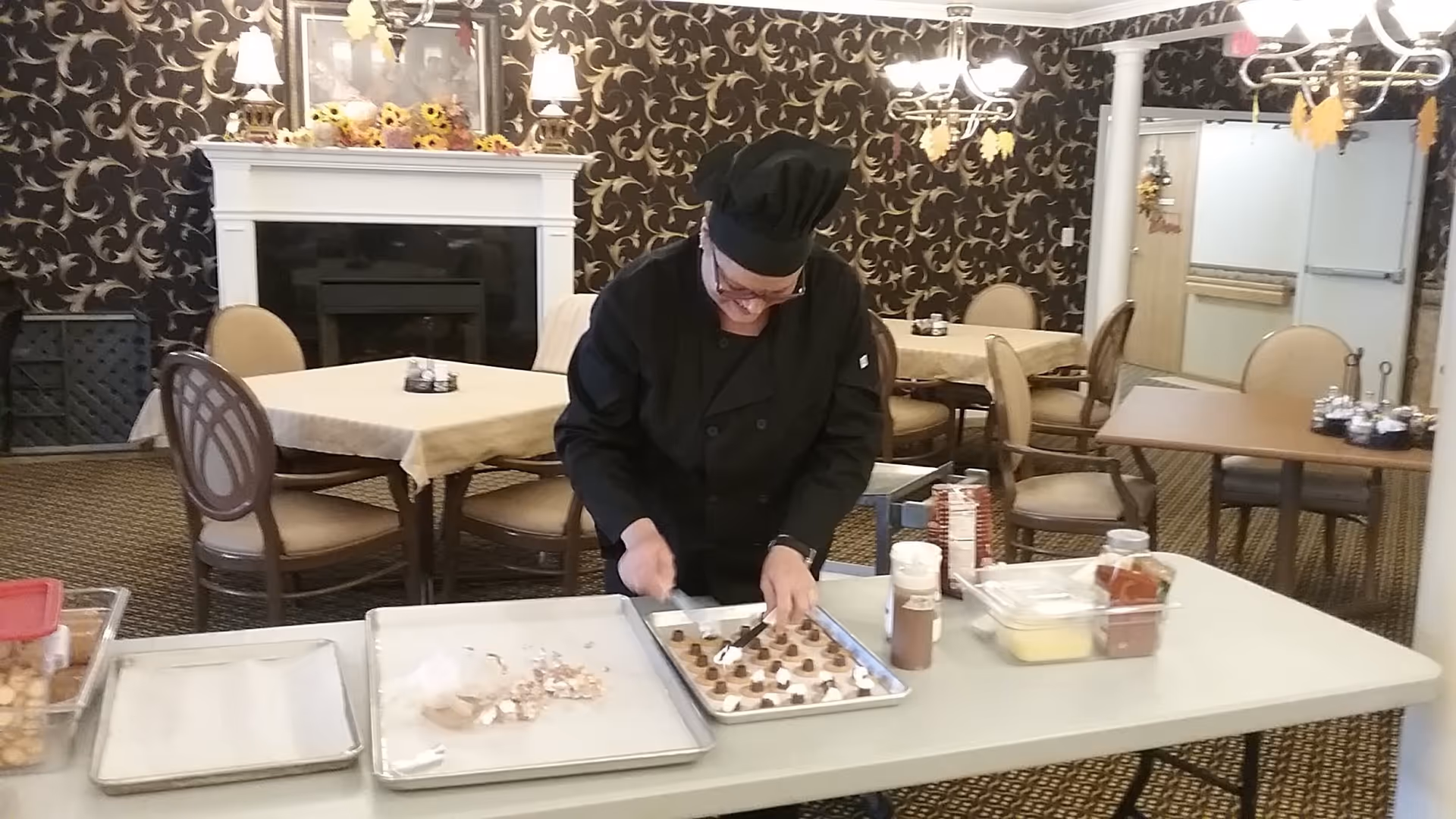 A chef in a black uniform prepares desserts on a tray at a folding table inside a decorated dining room with tables and chairs.