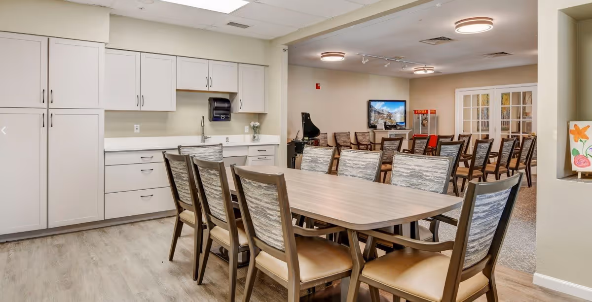 A spacious dining area in a senior living facility with a long wooden table surrounded by cushioned chairs. In the background, there is a kitchenette with white cabinets and a sink, and a seating area with additional chairs facing a wall-mounted TV. The room is well-lit with ceiling lights and has a clean, modern design.