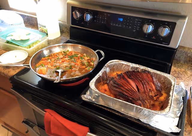 A kitchen stovetop with a skillet of mixed vegetables and a foil pan holding a glazed ham beside the oven controls.