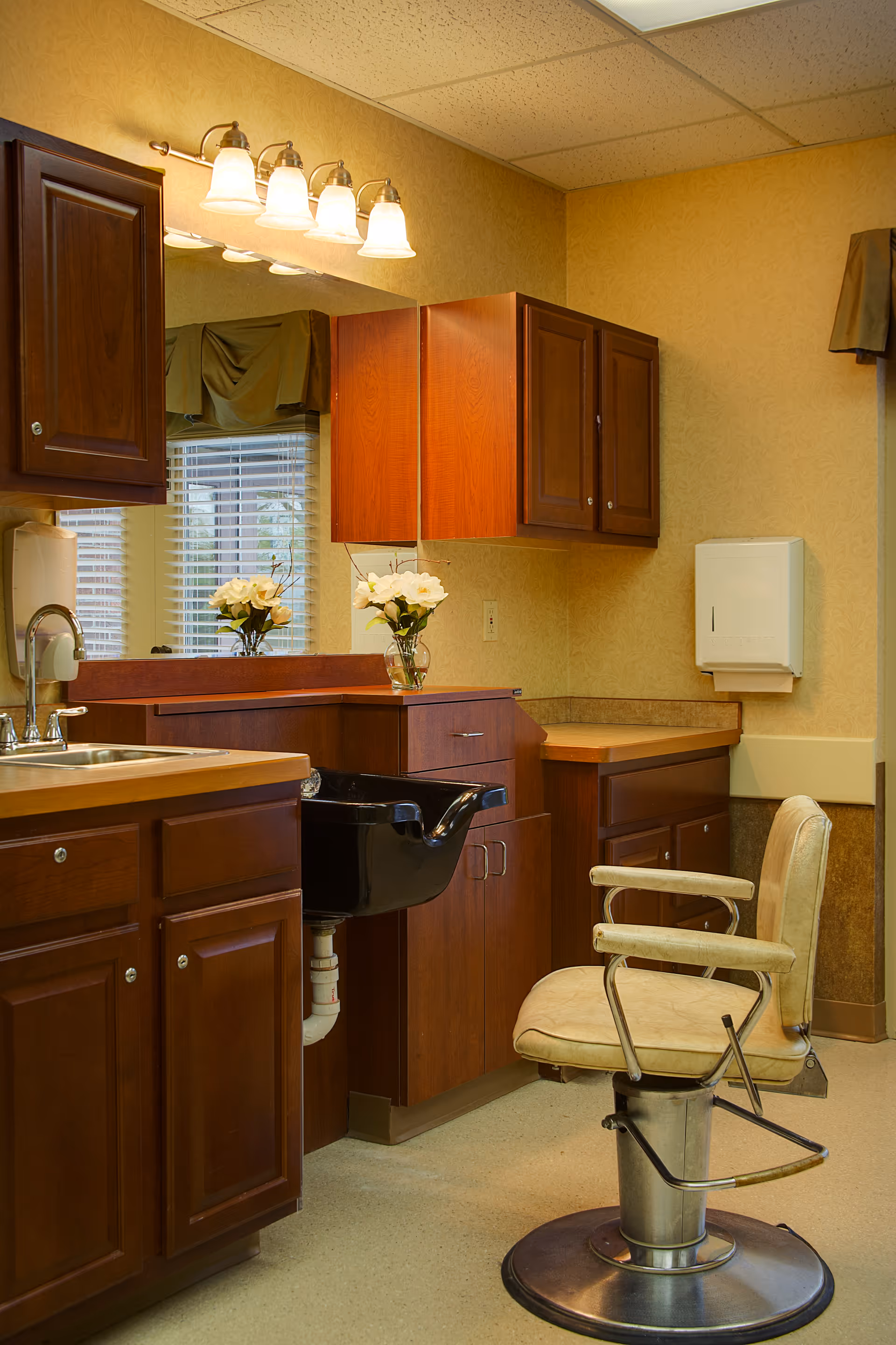 Interior view of a salon area in a senior living facility featuring a beige salon chair with metal armrests and footrest, a black wash basin, wooden cabinets, a countertop with a vase of white flowers, a large mirror, and a window with blinds and a valance.