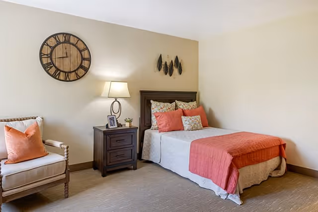 A cozy bedroom with a bed dressed in white and coral bedding, a wooden nightstand with a lamp and small decor items, a beige armchair with a coral pillow, a large round wall clock, and decorative wall art above the bed.