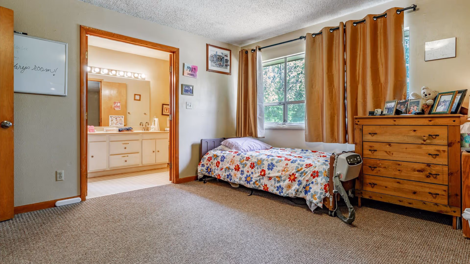A bedroom with a single bed covered in a colorful floral bedspread, positioned against a wall with a window that has brown curtains. Next to the bed is a wooden dresser with framed photos and a teddy bear on top. The room has beige walls and carpeted floor. An open doorway leads to a bathroom with a large mirror and vanity with multiple drawers and cabinets.