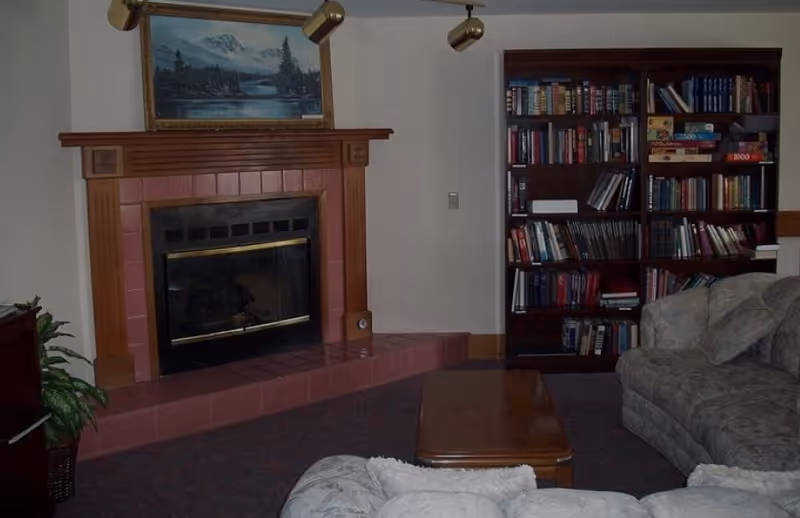 A cozy living room with a fireplace featuring a wooden mantel and a landscape painting above it. To the right of the fireplace is a tall bookshelf filled with books and board games. In front of the fireplace are two cushioned sofas and a wooden coffee table. A potted plant is visible to the left side of the fireplace.