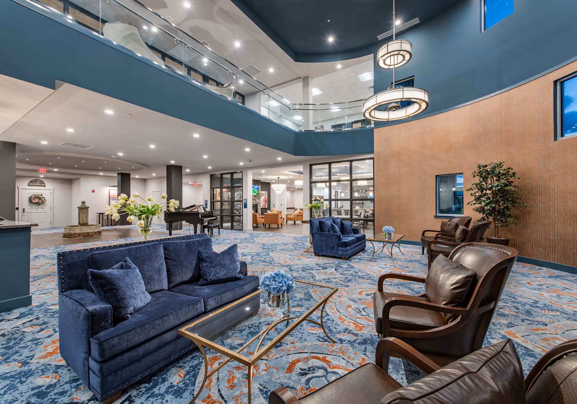 Spacious and modern senior living facility common area with blue velvet sofas, brown leather chairs, glass coffee tables with floral centerpieces, a grand piano, and a two-story ceiling with hanging circular light fixtures. The area features a patterned blue and orange carpet, large windows, and a balcony overlooking the space.