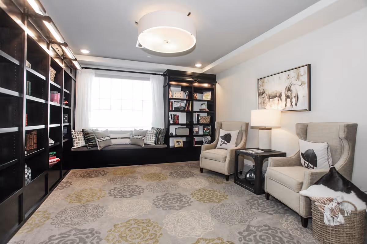 A cozy living room with a large window seat surrounded by built-in dark wood bookshelves filled with books and decorative items. Two beige armchairs with patterned pillows flank a small side table with a lamp. A basket with a blanket is placed next to one chair. The room has a patterned carpet and a large ceiling light fixture, with a framed artwork of animals on the wall.