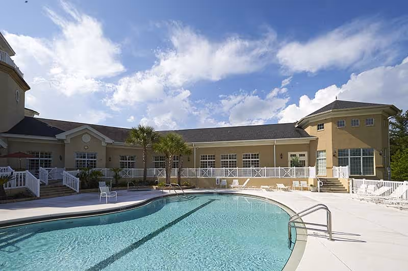 Outdoor swimming pool and sun deck with loungers in front of a low-rise senior living building under a blue sky.