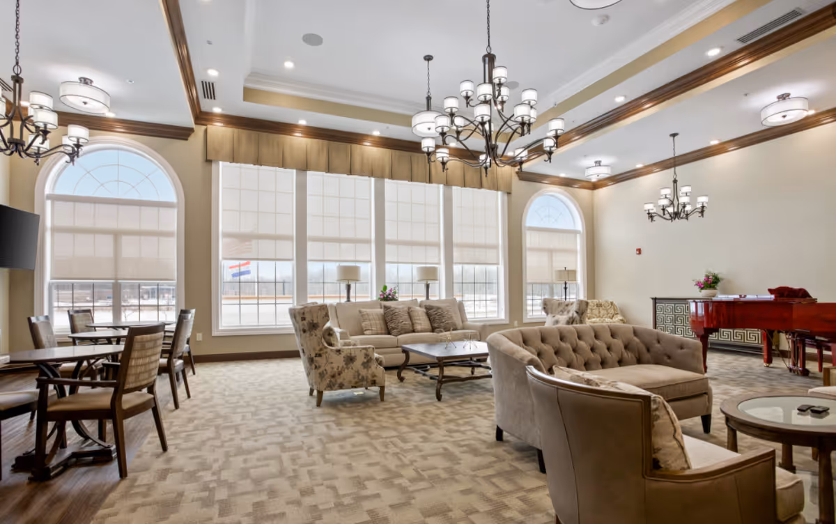 A spacious and well-lit senior living common area with large windows covered by white blinds, beige walls, and carpeted floors. The room features multiple seating arrangements including beige and floral upholstered sofas and armchairs, wooden tables, and a red grand piano. Elegant chandeliers hang from the ceiling, and there are table lamps and decorative plants placed around the room.
