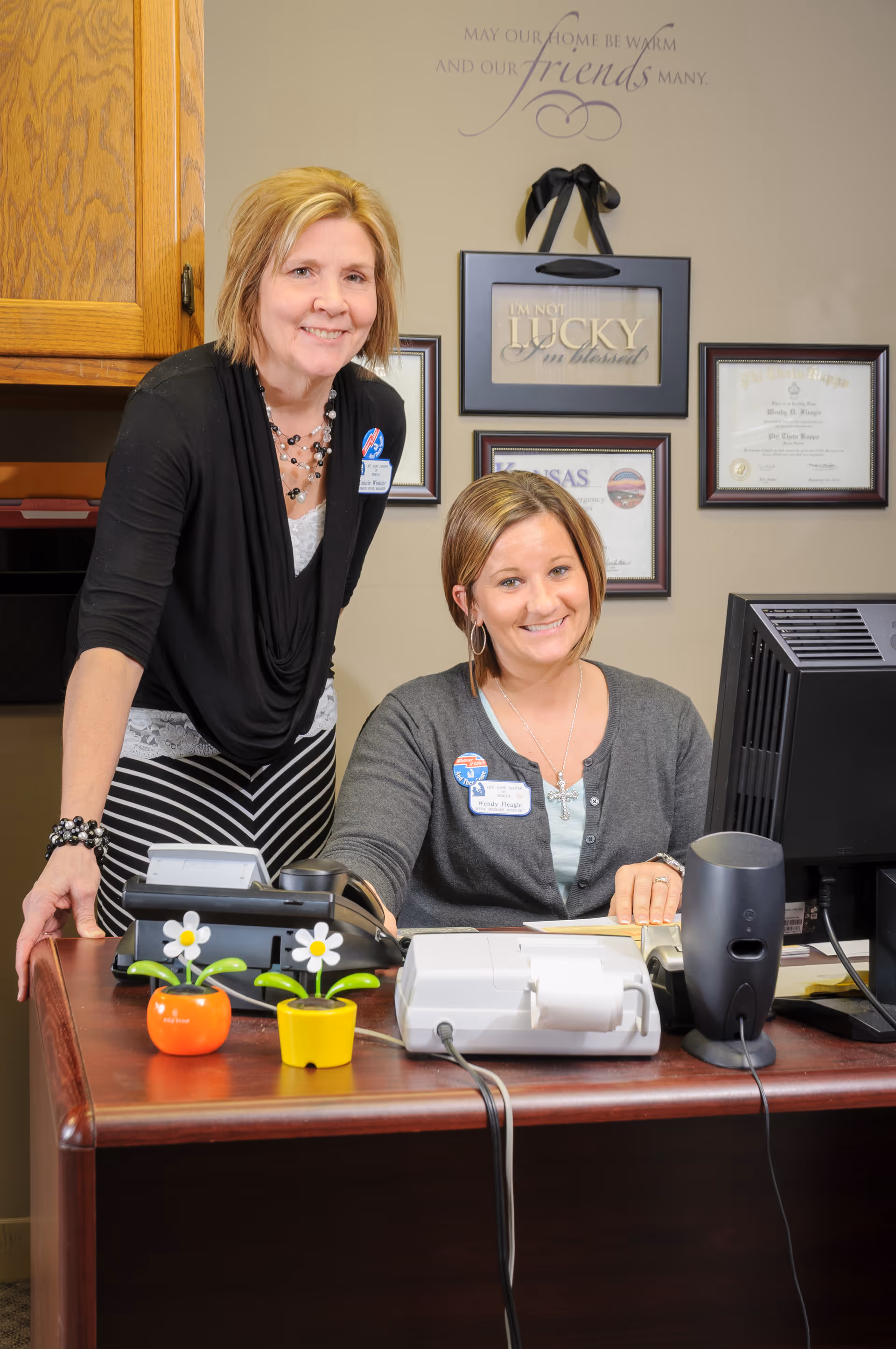 Two women smiling at a desk in an office setting. One woman is seated at the desk with a computer, phone, and office equipment, while the other woman stands beside her. The wall behind them has framed certificates and a decorative sign that reads 'I'm not lucky, I'm blessed.'