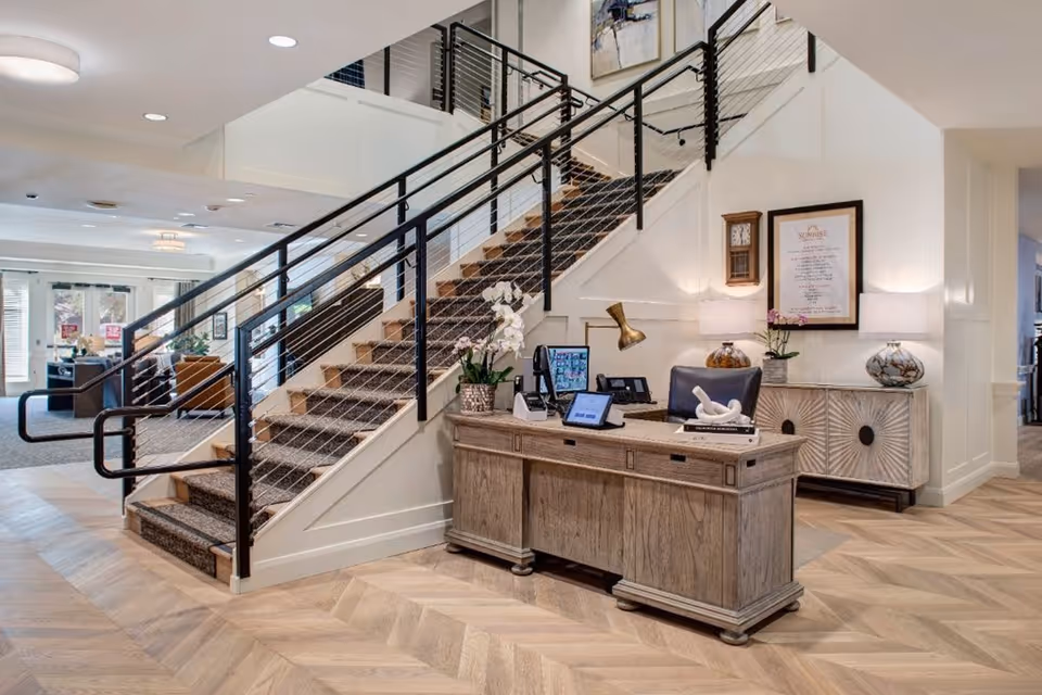 Interior view of a senior living facility lobby featuring a wooden reception desk with a computer, phone, and decorative items. Behind the desk is a staircase with a black metal railing and carpeted steps. The area is well-lit with ceiling lights and lamps on a sideboard against the wall. The flooring is light wood with a herringbone pattern.