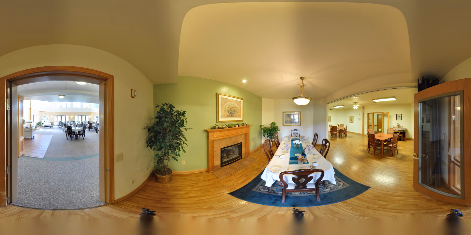 A spacious dining area in a senior living facility featuring a long dining table set with plates and napkins on a blue rug. The room has wooden flooring, a fireplace with a framed picture above it, and a potted plant. Adjacent rooms with additional tables and chairs are visible through open doorways.