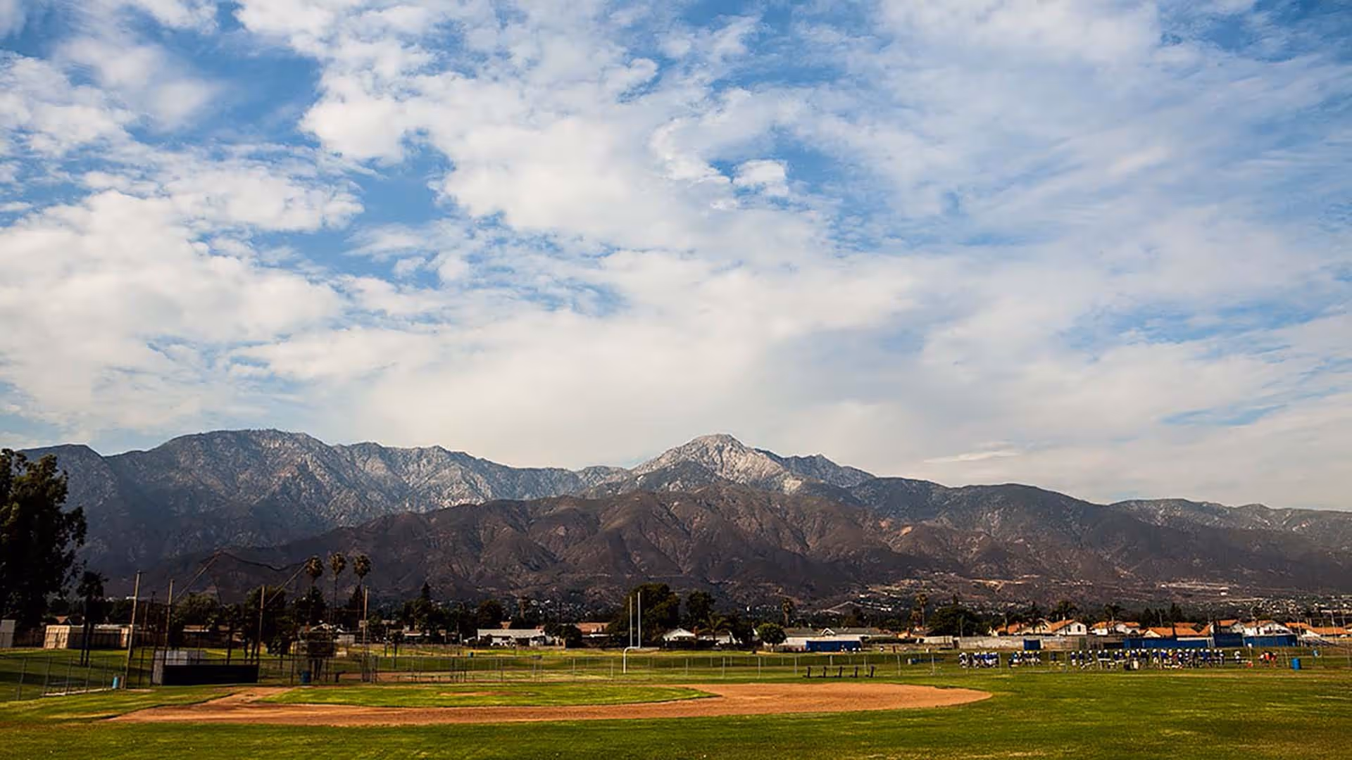 A wide view of a baseball field with green grass and a dirt infield, set against a backdrop of tall mountains under a partly cloudy blue sky. There are some trees, houses, and sports equipment visible in the distance.