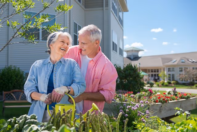 An elderly couple gardening together outdoors near a senior living facility. The woman is holding a yellow watering can and smiling, while the man stands behind her with his arms around her, both looking happy. There are green plants and flowers in the foreground and buildings in the background under a blue sky with some clouds.